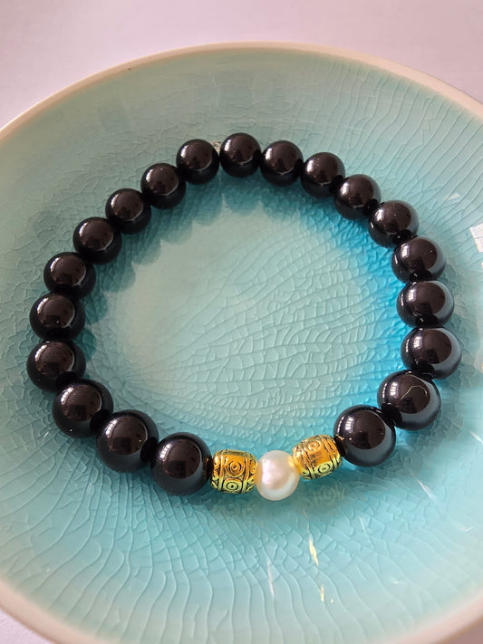 Black beaded bracelet with a gold and pearl charm on a textured blue plate.