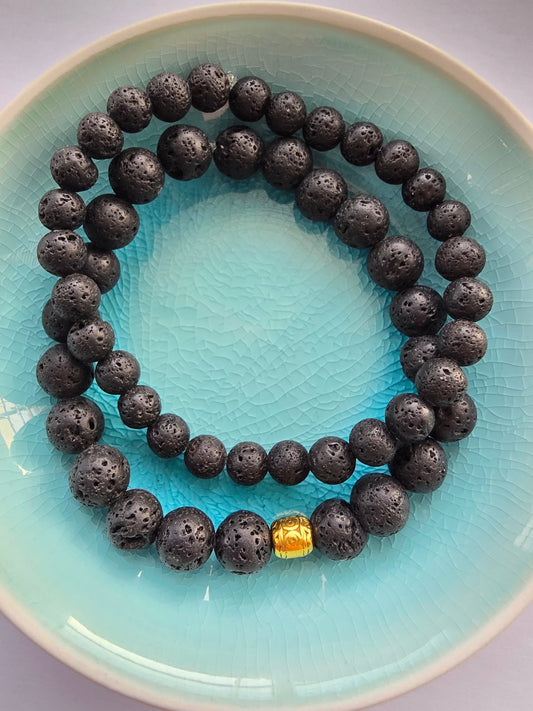 Pair of black lava stone beaded bracelets with a gold Tibetan bead accent on a light blue plate