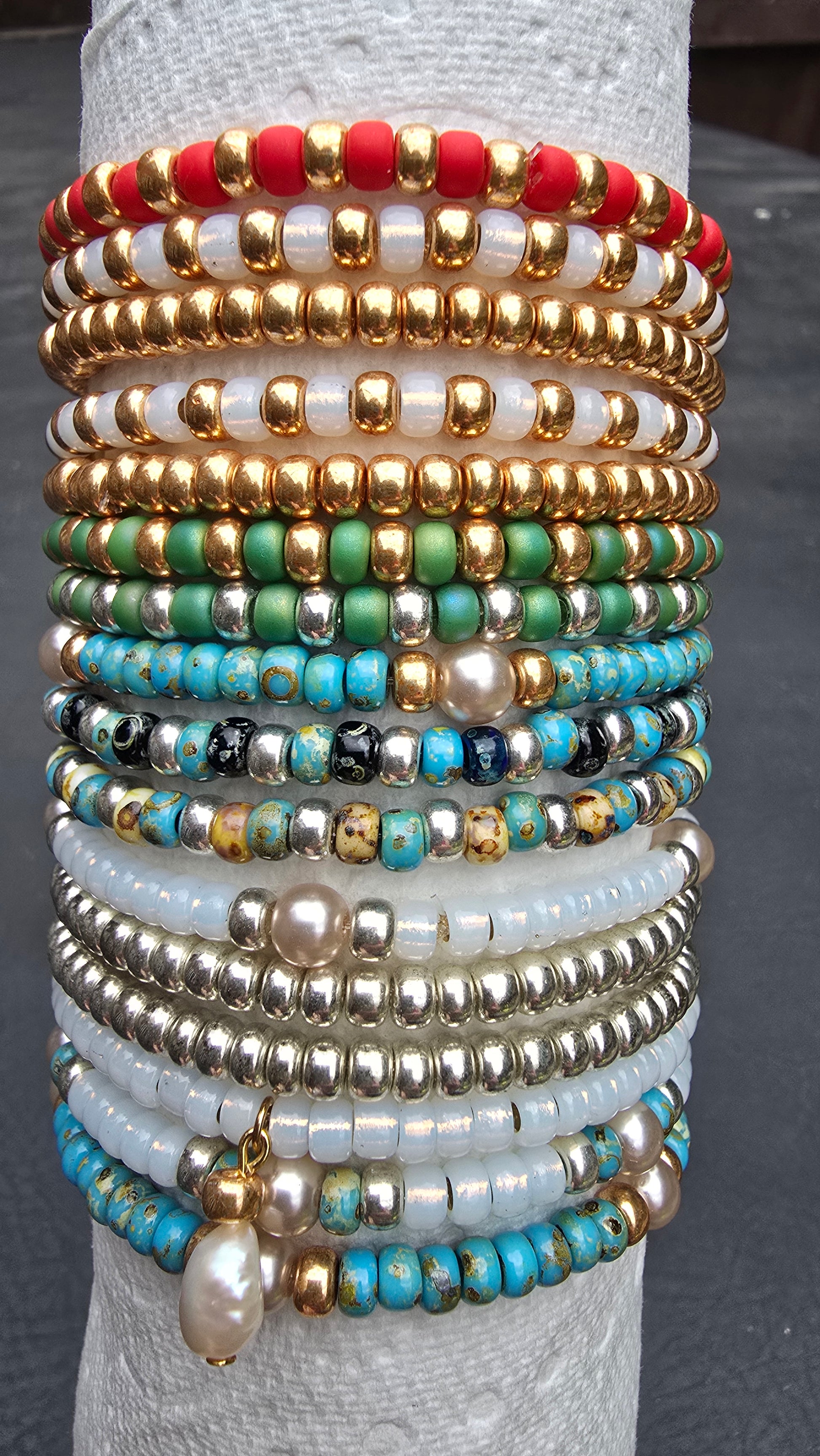 A stack of multiple beaded bracelets. The bracelets feature various bead combinations including gold beads, turquoise, white, red, green, and faux turquoise beads, with one larger white pearl charm hanging at the bottom.
