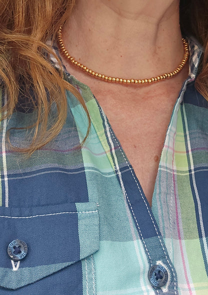 A close-up of a person wearing a delicate gold beaded chain necklace over a blue and green plaid button-up shirt.