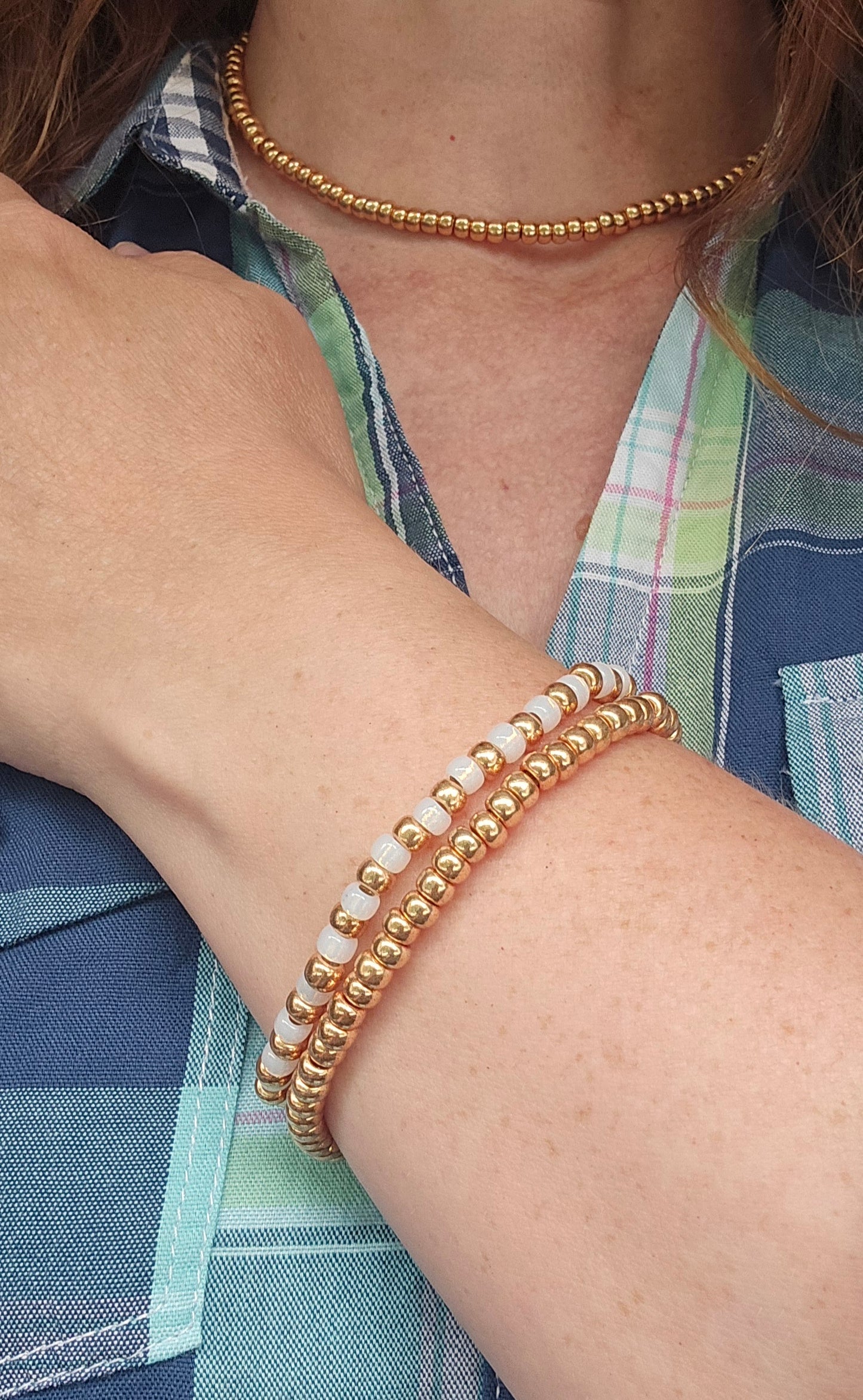 A close-up of a person's wrist wearing two beaded bracelets: two gold-toned beaded bracelets and one white beaded bracelet, with a matching gold beaded choker necklace also visible.
