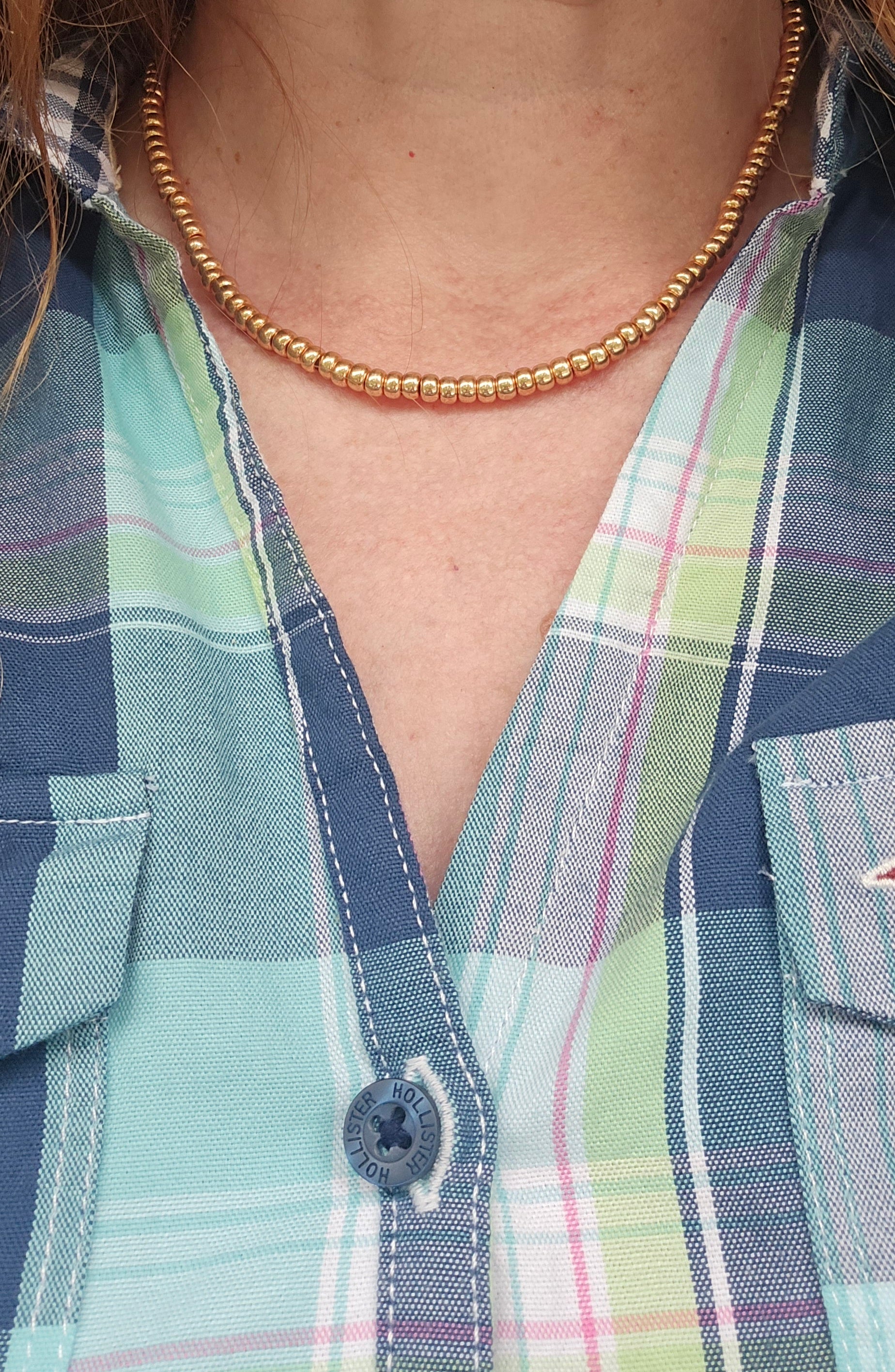 A close-up of a person wearing a gold beaded necklace over a plaid button-up shirt in shades of blue, green, and white.