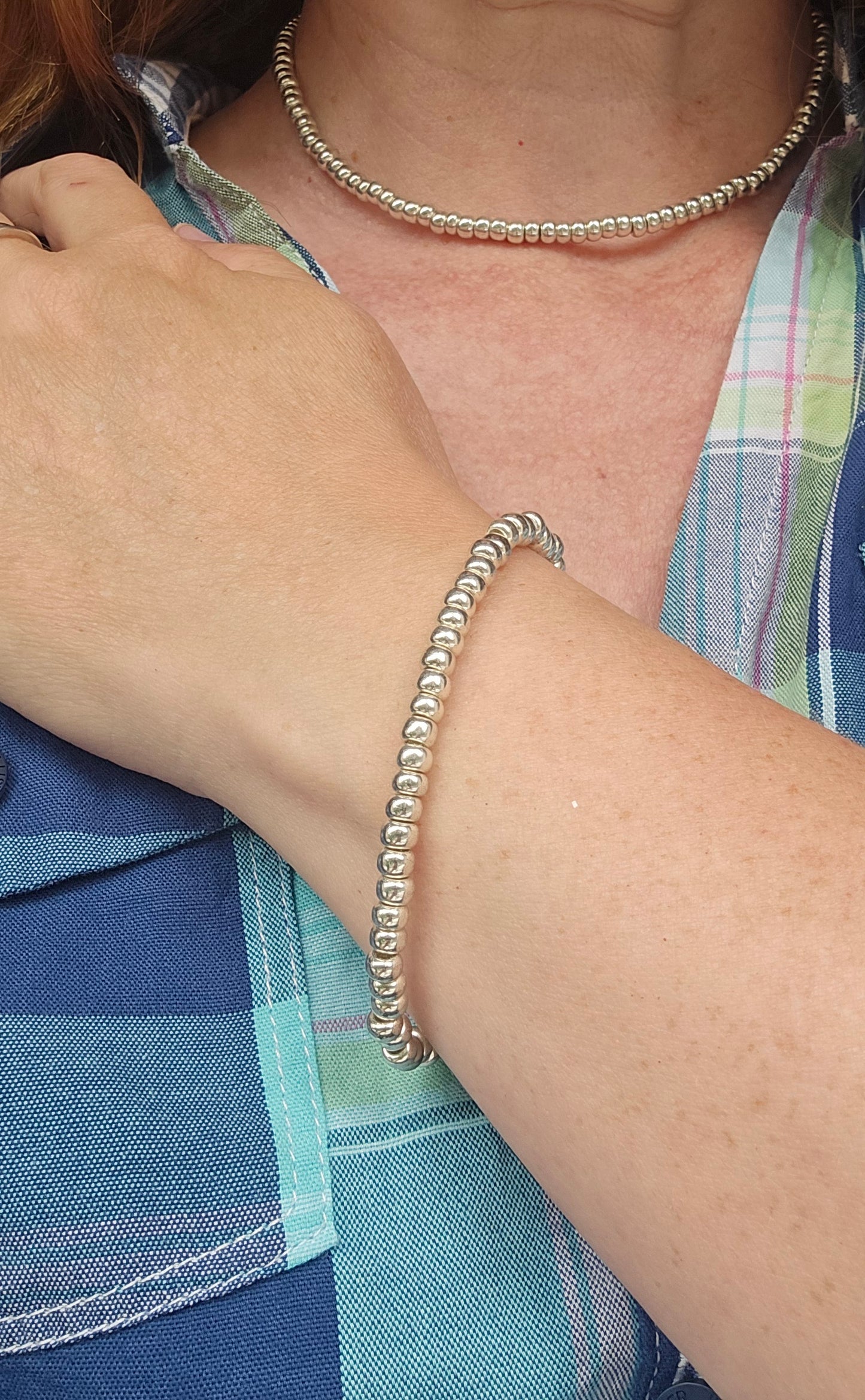 A close-up of a person's wrist wearing a silver beaded stretch bracelet, with a matching silver beaded choker necklace also visible against a blue and green plaid shirt.