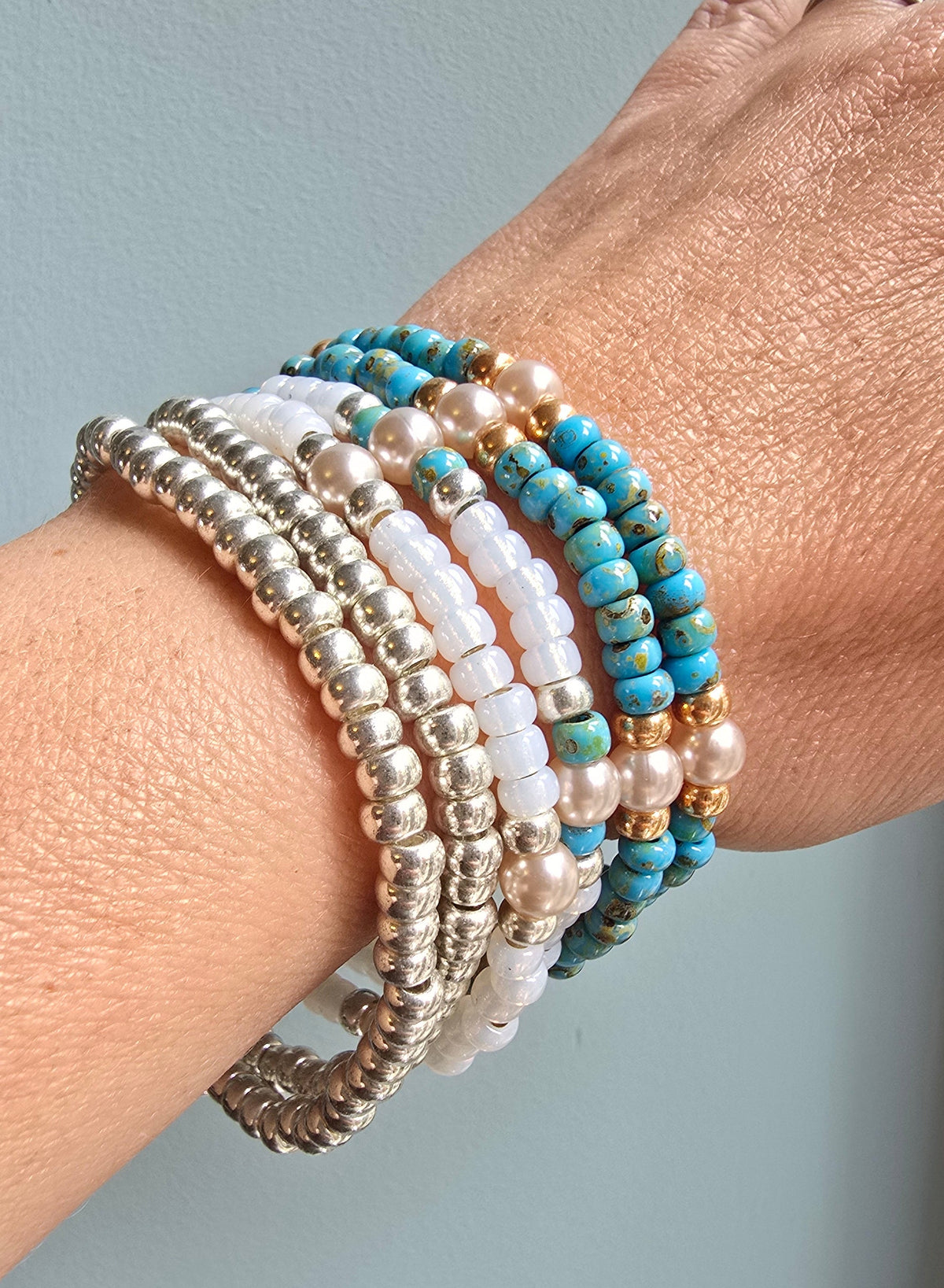 A close-up of a person's wrist wearing a stack of beaded bracelets, including silver-toned beads, white pearl-like beads, and turquoise blue beads with gold-toned accents.