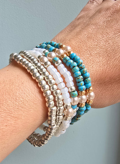 A close-up of a person's wrist wearing a stack of beaded bracelets, including silver-toned beads, white pearl-like beads, and turquoise blue beads with gold-toned accents.