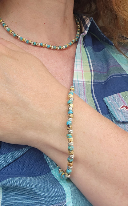 A close-up lifestyle shot of a person wearing a matching beaded necklace and bracelet. The jewelry features turquoise blue beads, light brown and silver beads.