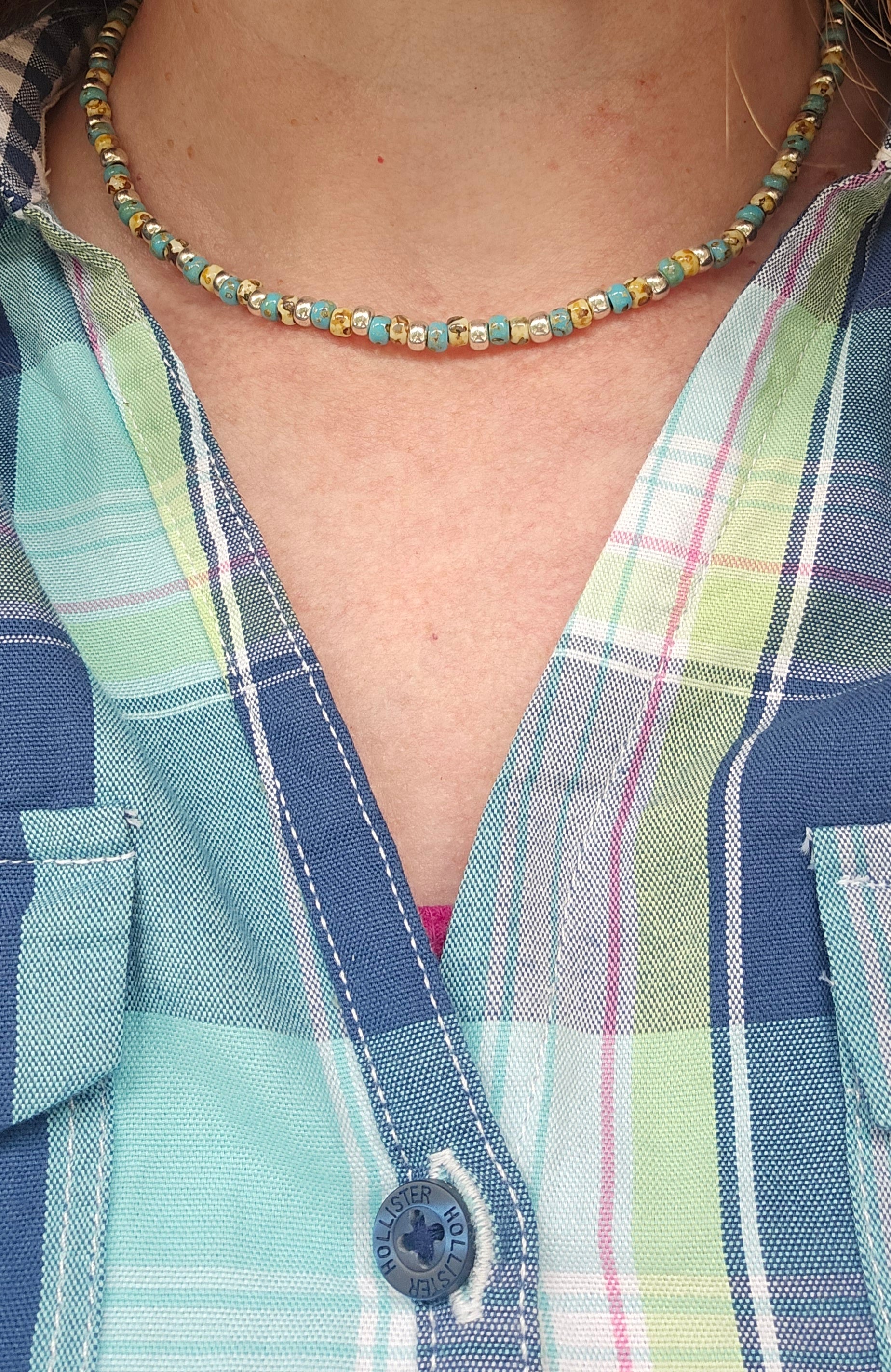 A close-up of a person wearing a beaded necklace with turquoise, gold, and brown beads, paired with a blue and green plaid button-up shirt.