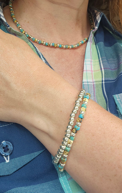 A close-up of a person wearing a beaded necklace and matching beaded bracelets. The jewellery features turquoise-blue, gold, and silver-toned beads, styled with a blue and green plaid shirt.