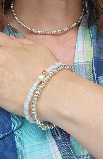 A close-up of a person's wrist wearing a stack of beaded bracelets, including a white pearl strand with a gold accent and a silver-toned beaded bracelet, along with a matching silver beaded choker necklace.