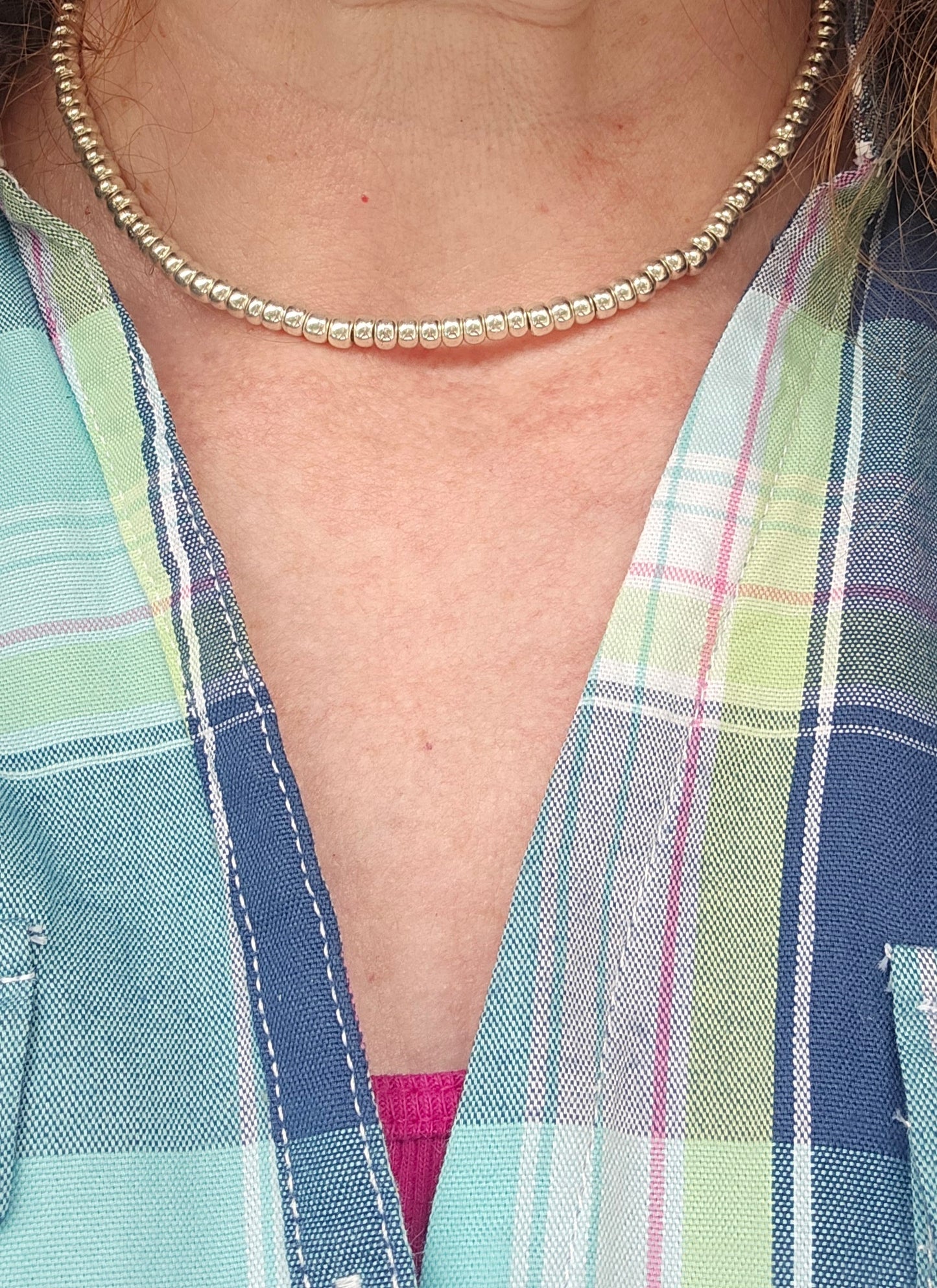 A close-up of a person wearing a delicate silver beaded necklace and a pastel plaid button-up shirt with blue, green, and pink tones.