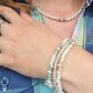A close-up of a person's neck and hand wearing multiple beaded jewelry pieces, including a pearl necklace, a multi-strand bracelet with white pearls, silver beads, and light blue accents, and a plaid shirt.