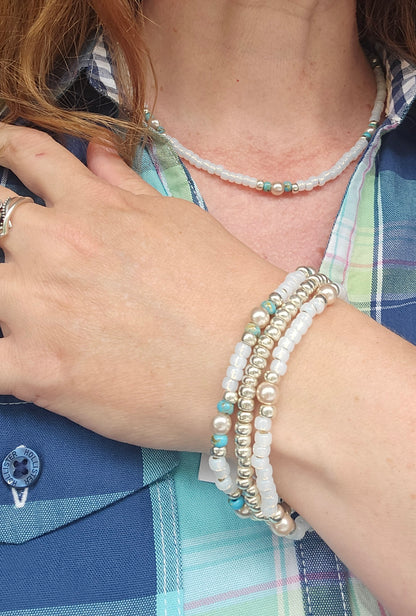 A close-up of a person's neck and hand wearing multiple beaded jewelry pieces, including a pearl necklace, a multi-strand bracelet with white pearls, silver beads, and light blue accents, and a plaid shirt.