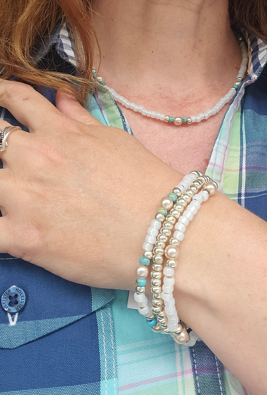 A close-up of a person's neck and hand wearing multiple beaded jewelry pieces, including a pearl necklace, a multi-strand bracelet with white pearls, silver beads, and light blue accents, and a plaid shirt.